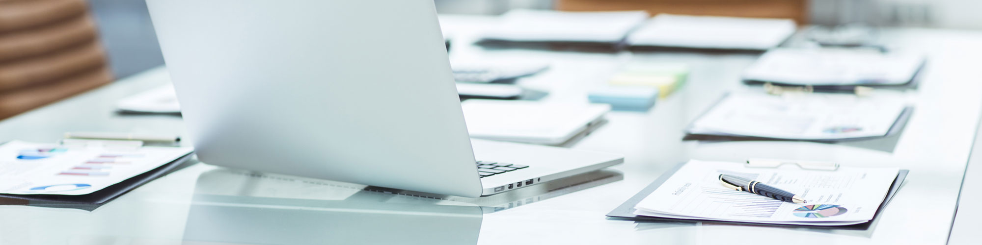 A laptop sits on a glass conference table surrounded by reports and charts, emphasizing a professional work environment and collaborative analysis.